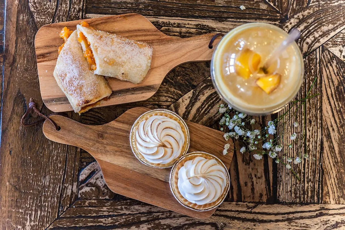 A wooden board with pastries and tarts beside two drinks.