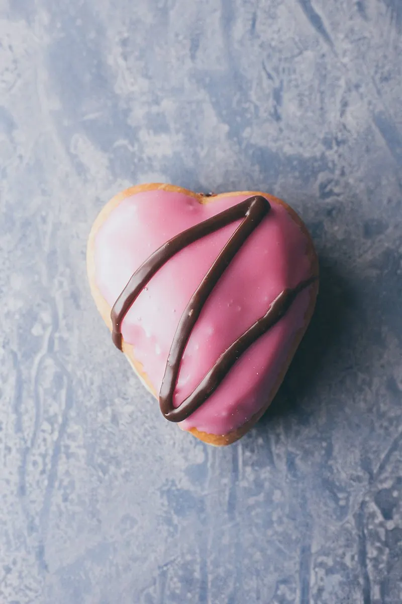 A heart shaped donut iced in pink with chocolate drizzle.